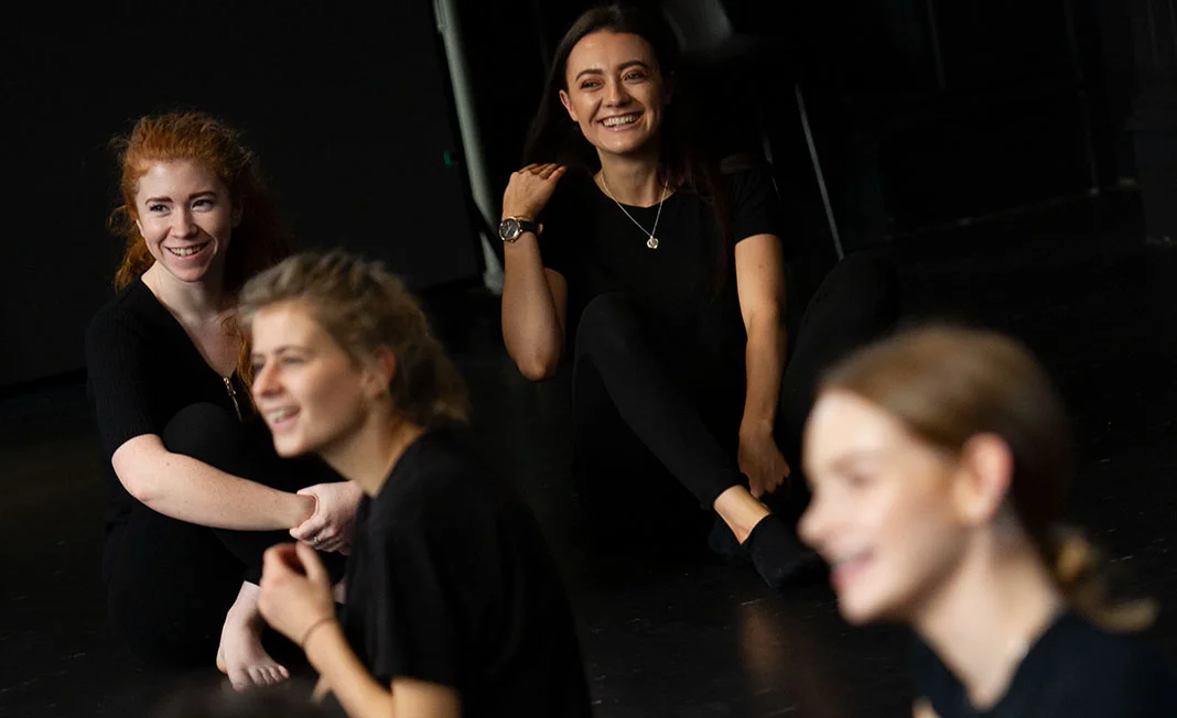 Arden learners in black clothing seated on a dark studio floor, with two in focus and two blurred in the foreground.