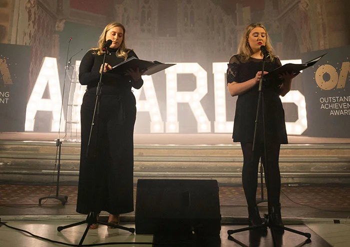 Two people standing at microphones on a stage, reading from scripts, with illuminated award lettering in the background.