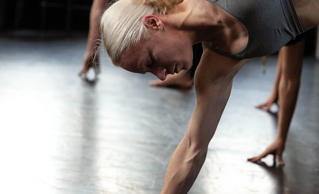 Person performing a floor-based exercise in a studio, supported on one arm, with other participants blurred in the background.