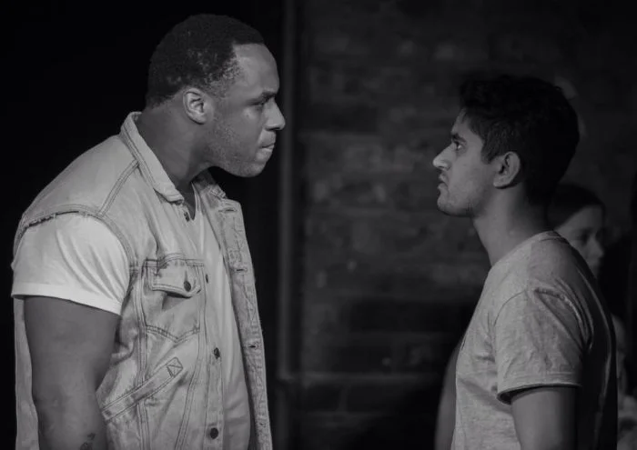 Two people standing face-to-face on a stage in a black-and-white scene, with another person partially visible in the background.